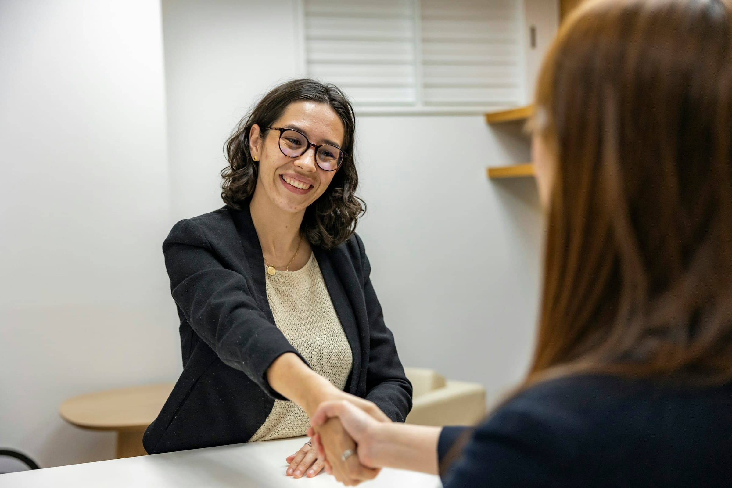 A smiling woman shakes hands across a desk during an interview