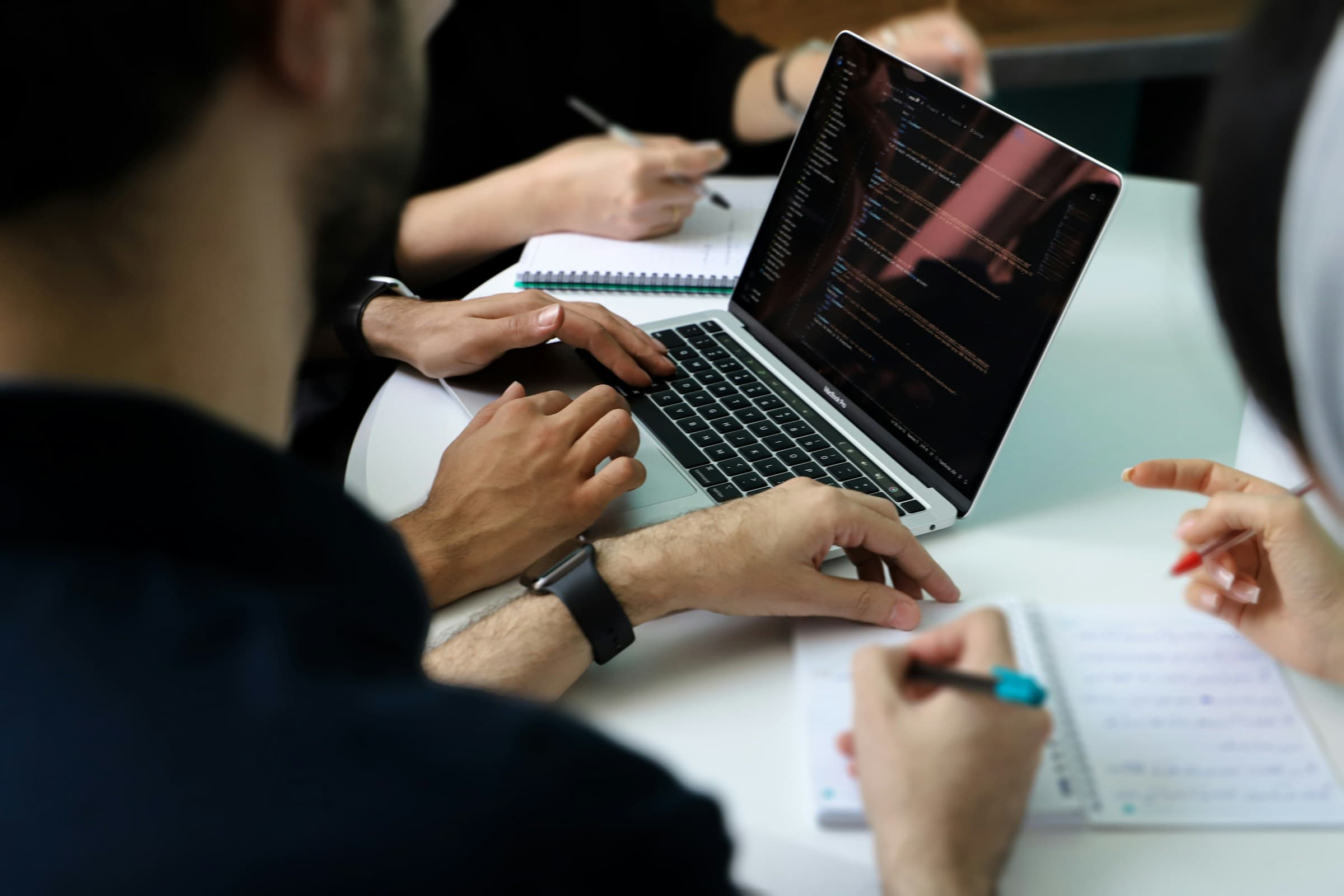 Hands on a laptop showing code, beside notes being written during a meeting