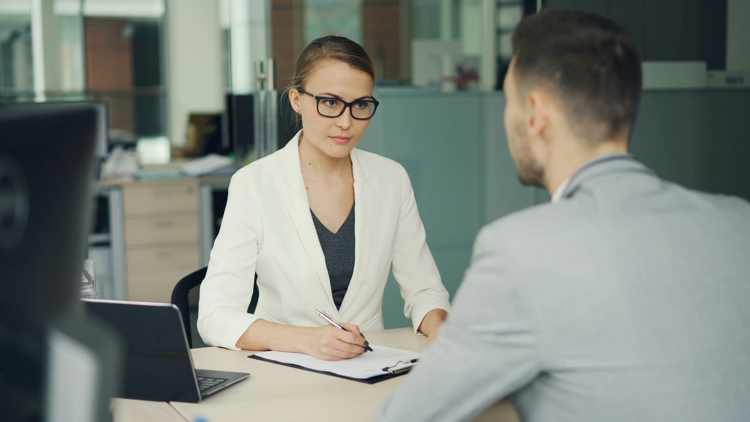 A woman in glasses takes notes while talking with a man across a desk
