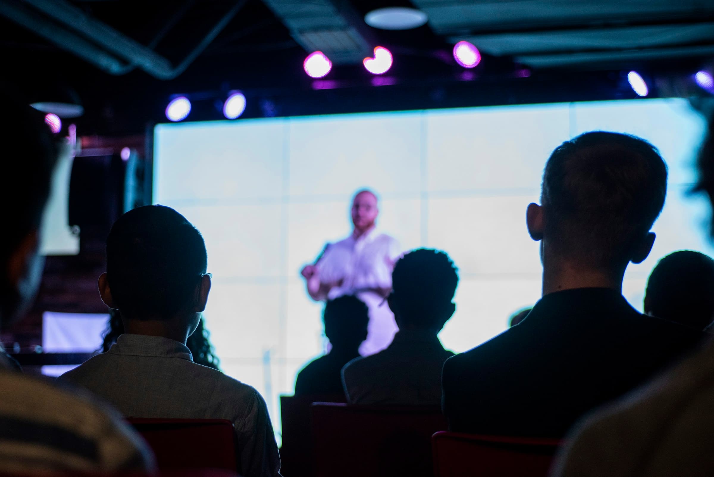 An audience in silhouette watching a speaker in front of a large, bright screen